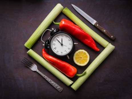 Place setting with a old style ringing alarm clock in the middle and a slice of orange. Flanked by 2 red chili peppers, surrounded in a square by 4 celery stalks. A fork and knife on the outside.
