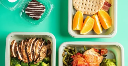 Three meal prep containers with grilled chicken, vegetables, sweet potato mash, oranges, flatbread with bacon, yogurt with berries, and a chocolate cookie