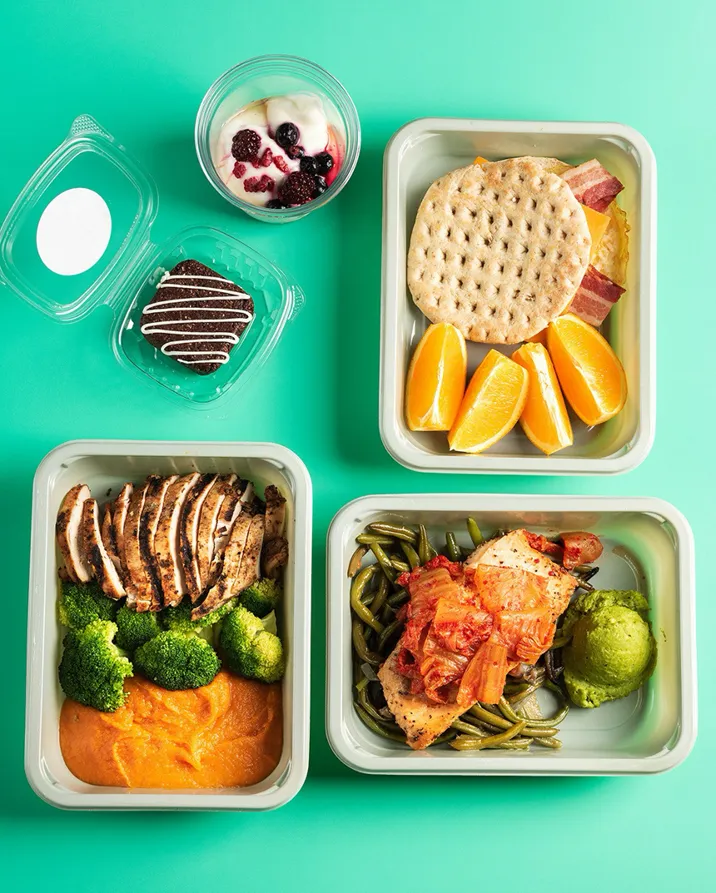 Three meal prep containers with grilled chicken, vegetables, sweet potato mash, oranges, flatbread with bacon, yogurt with berries, and a chocolate cookie