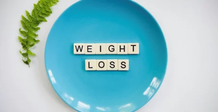 A blue plate with the words WEIGHT LOSS formed by letter tiles, and a green fern leaf beside the plate