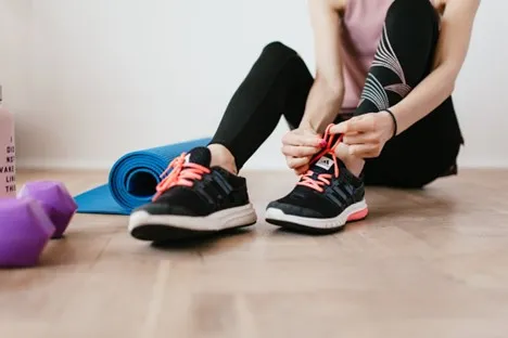 A person sits on the floor tying shoelaces on black athletic shoes with pink laces