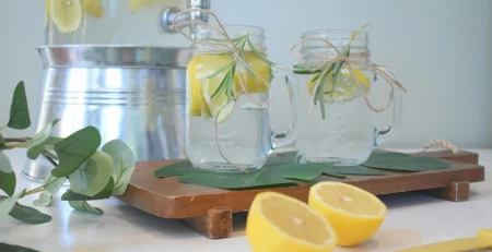 A refreshing beverage setup with two mason jars containing lemon slices and rosemary sprigs, tied with twine, and a large glass drink dispenser of lemon water in the background