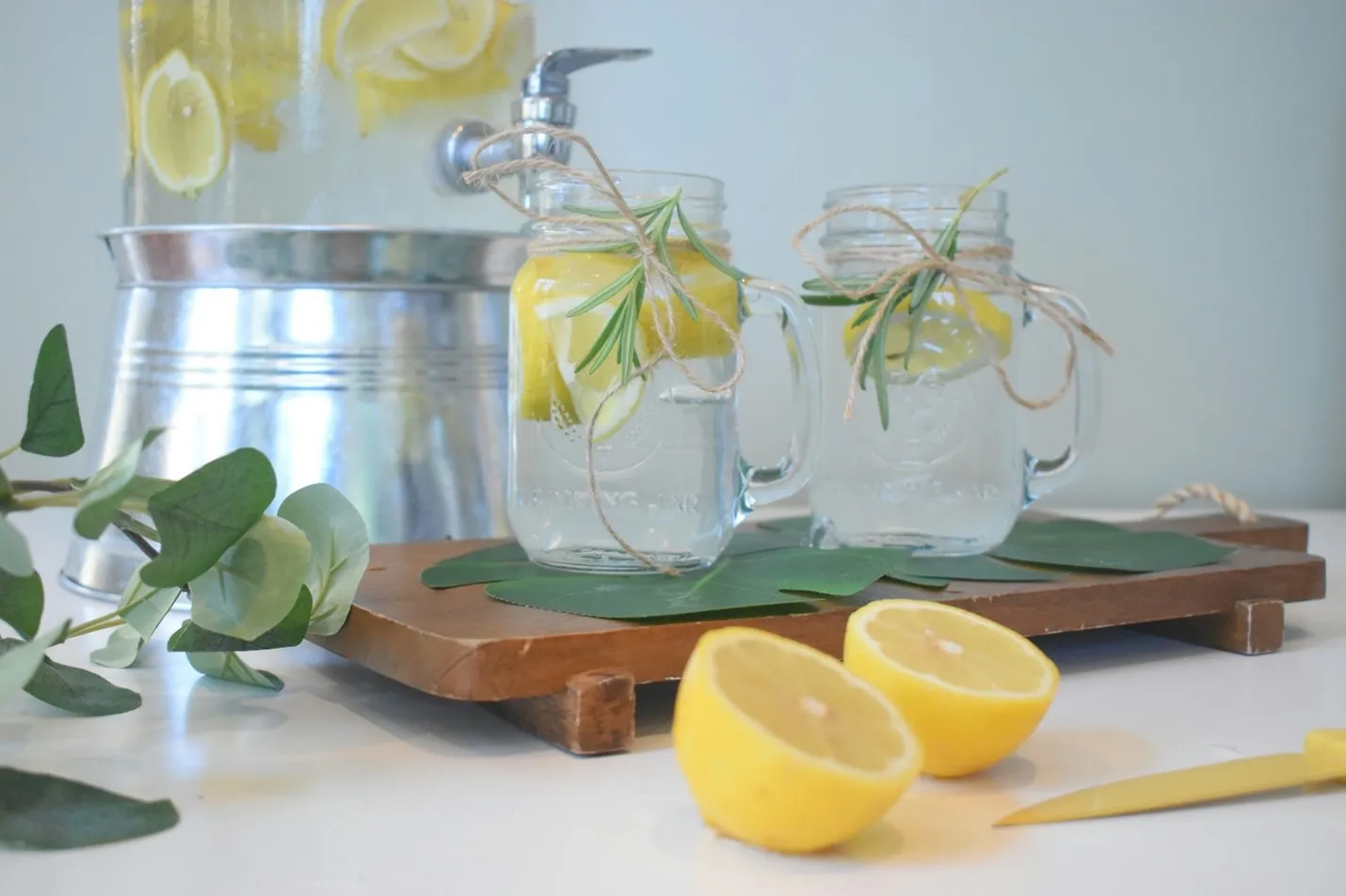 A refreshing beverage setup with two mason jars containing lemon slices and rosemary sprigs, tied with twine, and a large glass drink dispenser of lemon water in the background