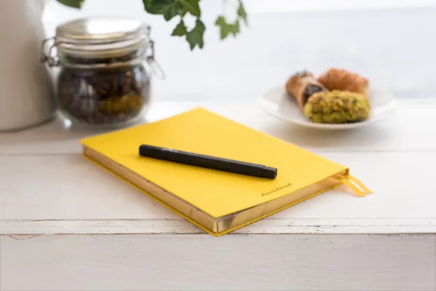 A yellow notebook with a black pen on it lies on a white wooden table. In the background, there is a jar, a small plant, and a plate with assorted pastries