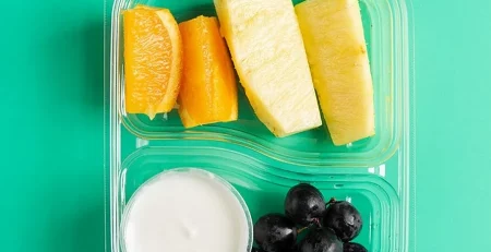 Fresh fruit slices with yogurt in a clear plastic container on a teal background.