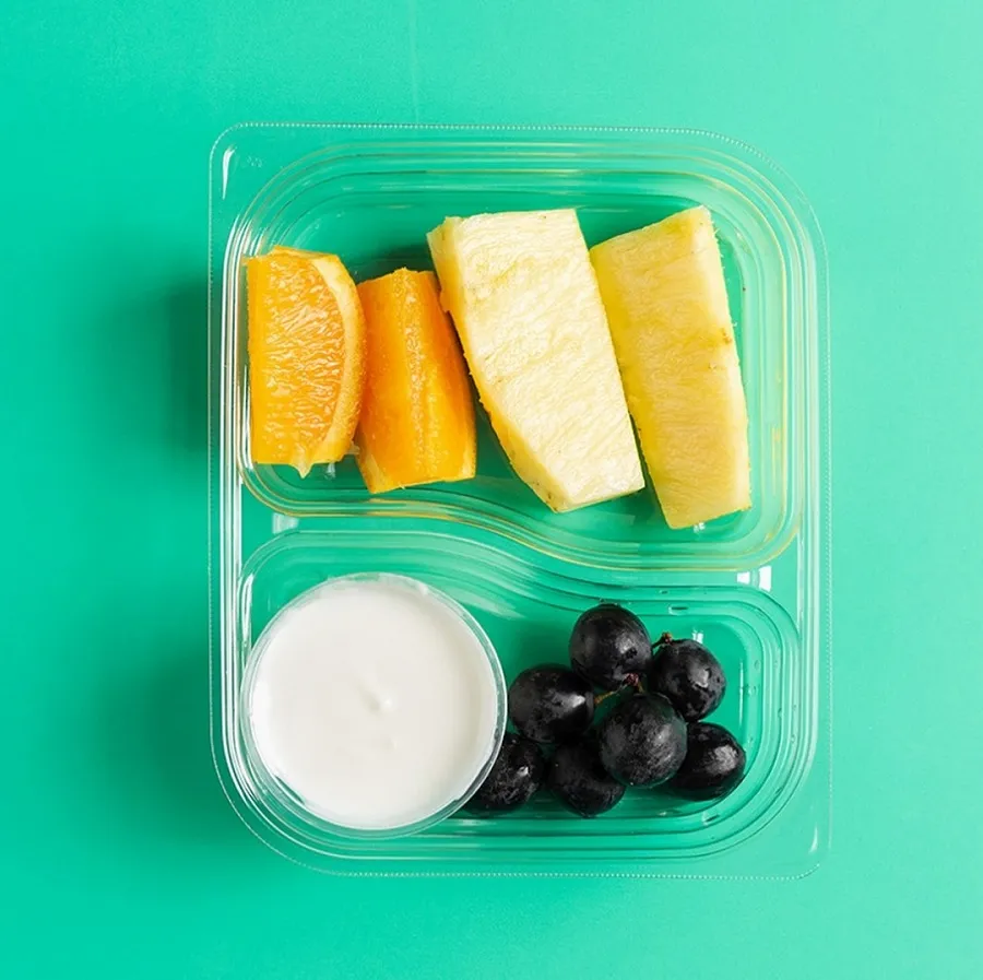 Fresh fruit slices with yogurt in a clear plastic container on a teal background.