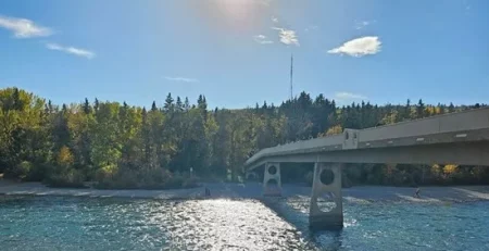 Sun over a river with a bridge and trees, person's feet in foreground