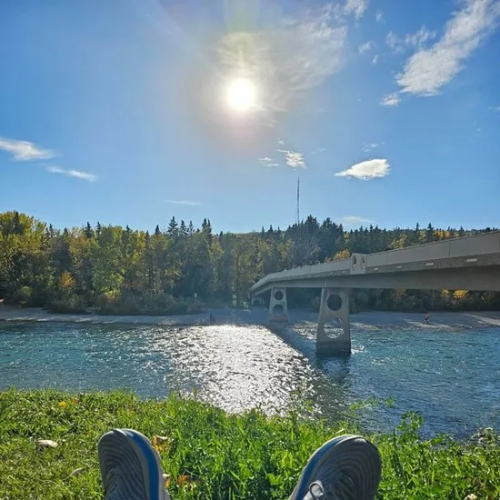 Sun over a river with a bridge and trees, person's feet in foreground