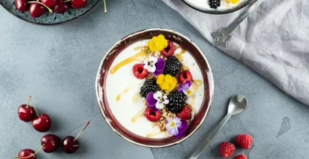 A bowl of yogurt with fresh berries and edible flowers, surrounded by cherries and raspberries