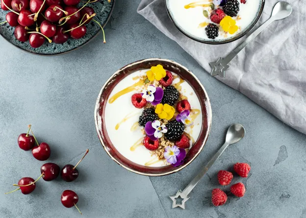 A bowl of yogurt with fresh berries and edible flowers, surrounded by cherries and raspberries